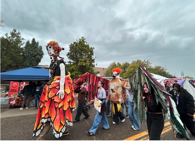 A colorful parade scene with large puppets and costumed participants walking past vendor tents under a cloudy sky.