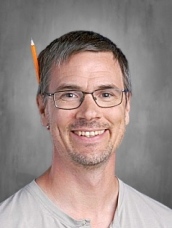 Hombre sonriente con gafas, pelo corto y un lápiz detrás de la oreja, vestido con una camisa clara sobre un fondo gris.