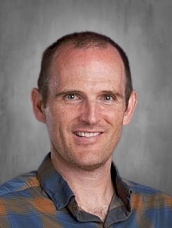 A smiling man with short hair, wearing a plaid shirt, against a gray background.