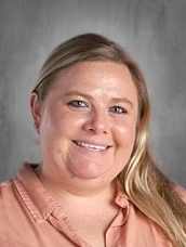 Smiling person with long hair, wearing a peach blouse, against a gray background.