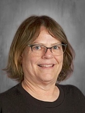 A person with glasses and shoulder-length hair, smiling at the camera against a gray background.
