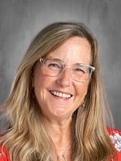 Woman with long blonde hair and glasses, smiling, wearing a red floral top against a gray background.