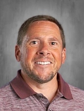 Man smiling, wearing a maroon collared shirt, with a gray background.