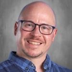 Smiling man with glasses and a blue plaid shirt against a gray background.