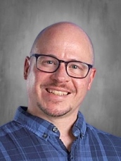 Hombre sonriente con gafas y una camisa a cuadros azules sobre un fondo gris.