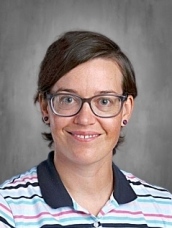 Person with glasses and short hair, wearing a striped shirt, smiling against a gray background.