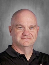 Bald man in a black polo shirt, smiling slightly, set against a gray background.