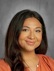 Person with long dark hair wearing an orange top, smiling at the camera against a gray background.