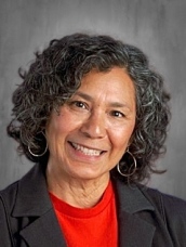 Smiling woman with curly hair, wearing a black blazer and red shirt against a gray background.