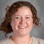 Smiling person with curly hair wearing a beige sweater and necklace against a gray background.