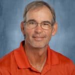 A smiling man with glasses and short gray hair wears an orange polo shirt against a blue background.