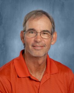A smiling man with glasses and short gray hair wears an orange polo shirt against a blue background.