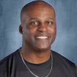 Smiling man with short hair wearing a black shirt and silver chain, against a blue background.