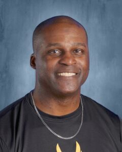 Smiling man with short hair wearing a black shirt and silver chain, against a blue background.
