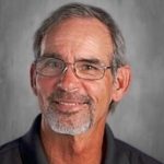 Man with glasses and a gray beard, wearing a dark shirt, smiling against a gray background.