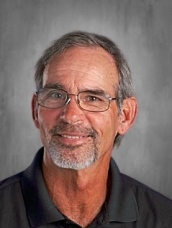 Man with glasses and a gray beard, wearing a dark shirt, smiling against a gray background.