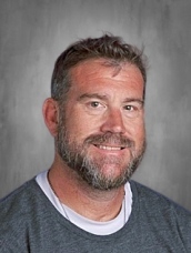 Man with short hair and a beard, wearing a gray shirt, smiling against a gray background.