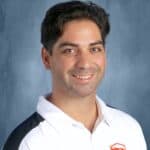 A smiling man with dark, curly hair wearing a white polo shirt with a volleyball logo, against a blue background.