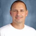 Smiling man with short hair wearing a white t-shirt with a volleyball graphic, set against a blue background.