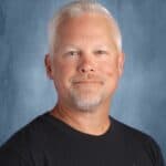 A smiling man with short white hair and a beard, wearing a black T-shirt, poses against a blue background.