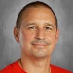 Man in a red shirt smiling at the camera against a gray background.