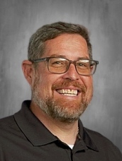 Man with short hair, beard, and glasses, wearing a dark collared shirt, smiling against a gray background.