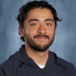 A man with curly hair and a beard smiles, wearing a dark hoodie with "Skyline" logo on it. Background is blue.