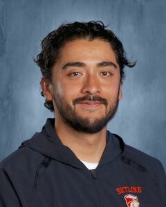 A man with curly hair and a beard smiles, wearing a dark hoodie with "Skyline" logo on it. Background is blue.
