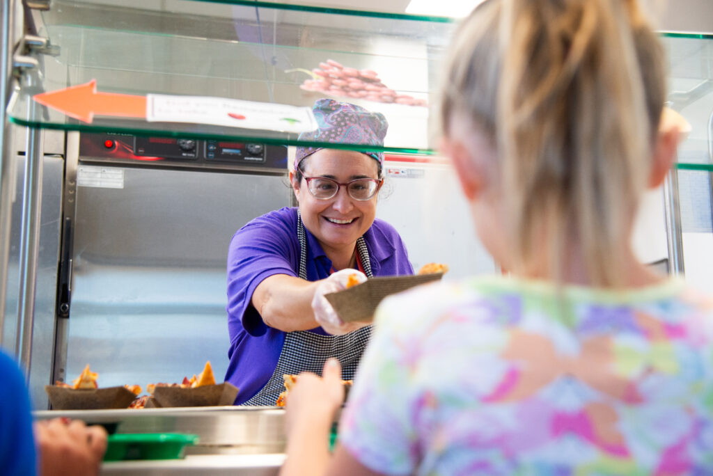 A smiling server hands food to a child at a cafeteria, with trays of food visible in the background.