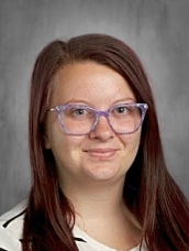 A person with long hair and glasses smiles slightly against a gray background.