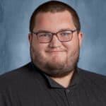 A young man with glasses and a beard smiles in front of a blue background, wearing a dark collared shirt.