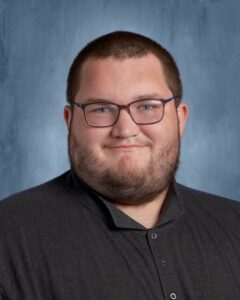 A young man with glasses and a beard smiles in front of a blue background, wearing a dark collared shirt.