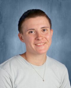 Smiling person with short hair wearing a light gray shirt and a necklace, against a blue background.