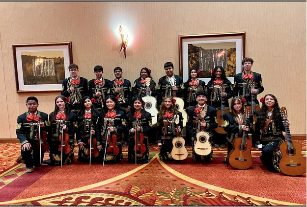 A group of 16 Mariachi student musicians in traditional attire, holding instruments, poses in a warmly lit indoor setting.