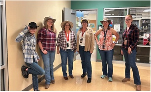 cinco mujeres y un estudiante vestidos con atuendos vaqueros, incluidos sombreros y camisas de cuadros, posan juntos en un pasillo muy iluminado.