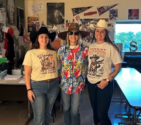Tres mujeres vestidas con trajes temáticos permanecen juntas en un aula decorada con carteles y recuerdos.