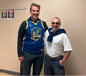 Dos hombres posan juntos en un pasillo; uno lleva una camiseta de baloncesto, el otro una camisa blanca con un jersey al cuello.