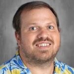 Smiling man with short hair wearing a colorful Hawaiian shirt against a plain background.