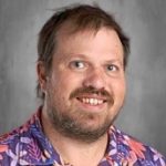A person with short hair and a beard, wearing a colorful floral shirt, smiles in front of a gray background.