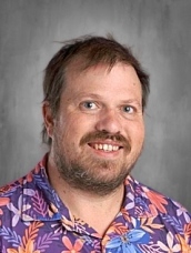 A person with short hair and a beard, wearing a colorful floral shirt, smiles in front of a gray background.