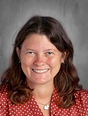 Smiling woman with long brown hair, wearing a red polka dot blouse, against a gray background.
