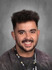 Hombre sonriente con el pelo corto y rizado, vestido con una camiseta oscura y un cordón, sobre un fondo gris.