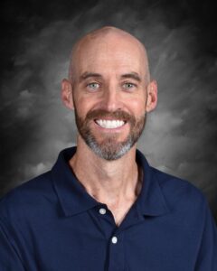 Smiling man with a bald head and beard, wearing a navy polo shirt, set against a dark gray background.