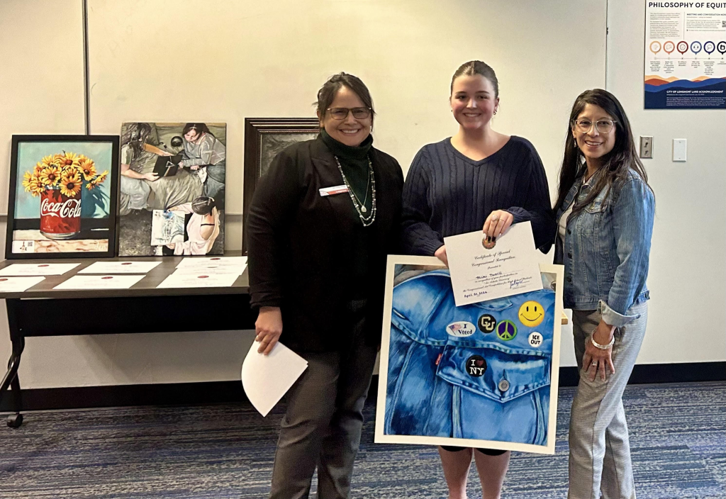 Three women stand together, holding a certificate and a colorful artwork of a denim jacket with various pins.