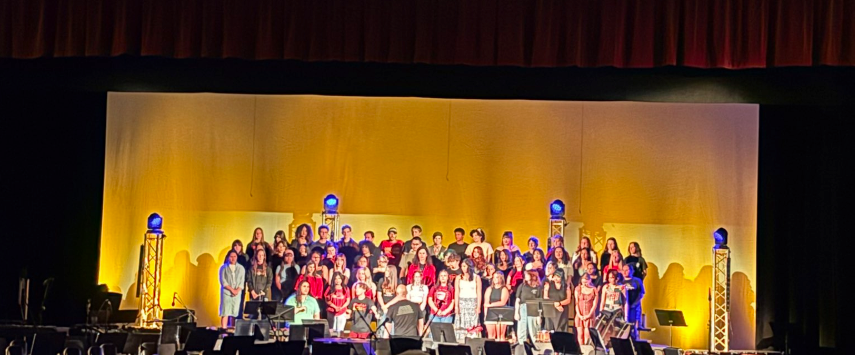 A large group of students performs on stage, with colorful lighting and musical instruments visible in the foreground.