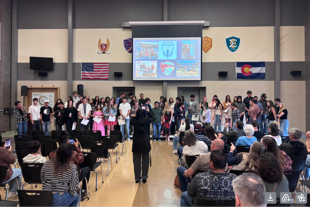 A group of students stands on stage during a ceremony, while an audience watches and takes photos.