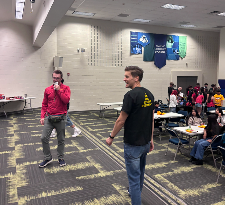 Two men interact in a large room filled with tables and students, with banners hanging on the walls.