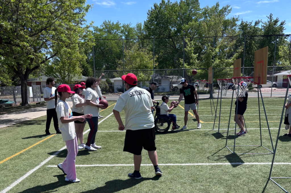A diverse group of children and adults play basketball on a sunny court, with one participant in a wheelchair.