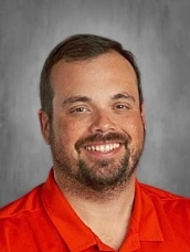 Man with short dark hair and beard, wearing an orange shirt, smiling against a gray background.