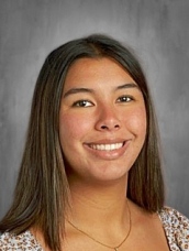 Smiling person with long hair, wearing a patterned top, against a gray background.